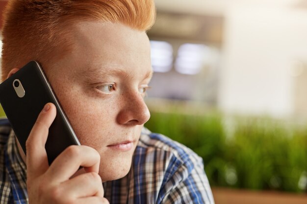 Zamyka w górę portreta modny facet z rudymi włosami ma piegi na jego twarzy opowiada przez telefon podczas gdy siedzący w restauraci patrzeje zamyślony na boku. Ludzie, moda, koncepcja technologii.