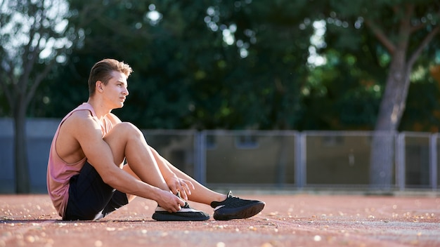 Zamyka W Górę Strzału Atleta Biegacza Młoda Samiec Rozciąga Jego Nogę Na Biegowym śladzie W Stadium, Przygotowywa Dla Pracującego Out. Kaukaski Mężczyzna ćwiczy Outdoors Być Ubranym Błękitną Odzież Sportową. Sport, Ludzie, Styl życia