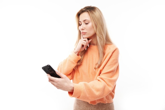 Zamyślona blond dziewczyna w swobodnym trzymaniu smartfona na białym tle studio