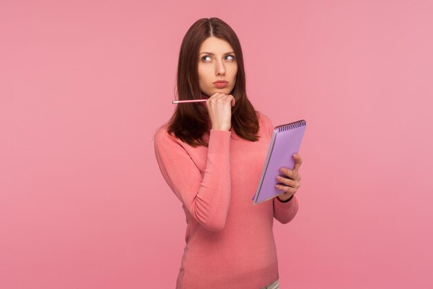 Zamyślona brunetka kobieta w różowym swetrze trzymająca notatnik i ołówek w dłoniach zastanawiająca się nad planami uruchomienia Ujęcie w studio na białym tle na różowym tle