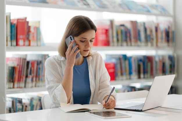 Zamyślona kobieta rozmawiająca przez telefon komórkowy, korzystająca z laptopa pracującego online, robiąca notatki w nowoczesnym biurze