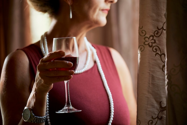 Zamyślona, melancholijna kobieta w średnim wieku trzymająca kieliszek wina i patrząca na zewnątrz przez okno