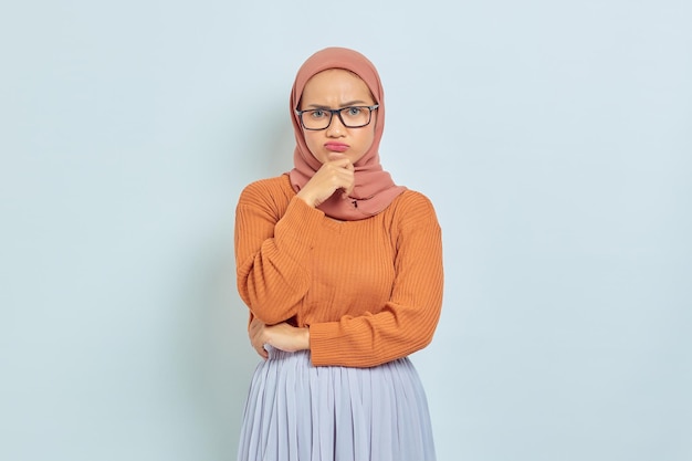 Zamyślona młoda azjatycka muzułmanka w brązowym swetrze i okularach trzymająca podbródek, myśląc o czymś na białym tle ludzie religijny styl życia koncepcja