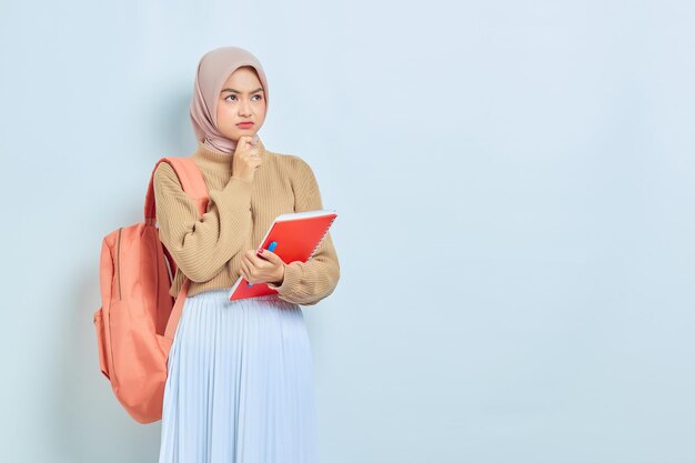 Zamyślona młoda azjatycka muzułmańska studentka w brązowym swetrze z plecakiem trzymająca książkę i myśląca o czymś na białym tle powrót do koncepcji szkoły