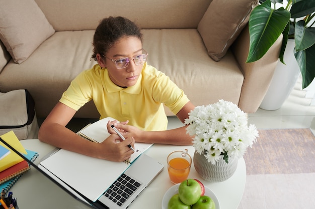 Zamyślona nastolatka rasy mieszanej w okularach siedzi przy stoliku do kawy z kwiatami w wazonie i korzysta z laptopa podczas robienia notatek na temat artykułu online