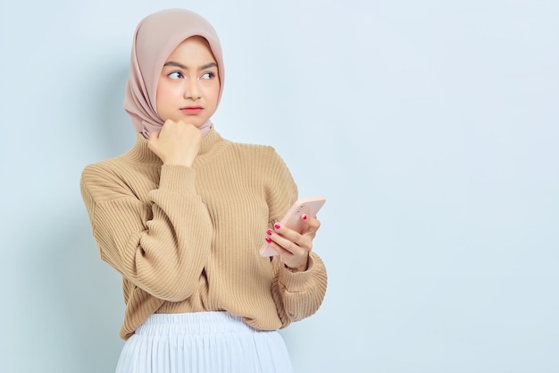 Zamyślona piękna azjatycka muzułmanka w brązowym swetrze trzymając telefon komórkowy wygląda poważnie myśląc o czymś izolowanym na białym tle Koncepcja religijnego stylu życia ludzi