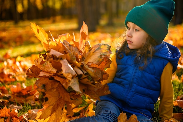 Zamyślona piękna dziewczynka w jasnych kolorowych ubraniach odwracająca wzrok, pozująca z żółtym bukietem suchych liści klonu siedząca wśród opadłych liści na jesiennym tle przyrody z miejscem na kopię