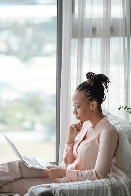 Zamyślona poważna przedsiębiorczyni w piżamie czytająca dokument na ekranie laptopa podczas pracy w domu