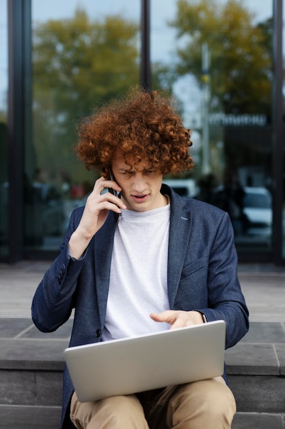 Zamyślony atrakcyjny mężczyzna używający laptopa rozmawiający na smartfonie dokonujący zamówienia Przystojny freelancer pracujący online na świeżym powietrzu