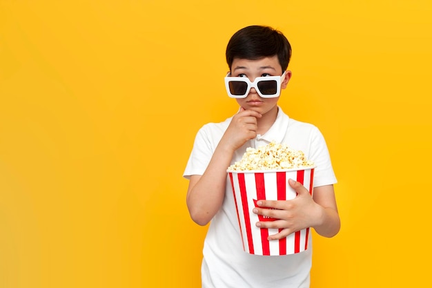 zamyślony azjatycki mały chłopiec w wieku dziesięciu lat w okularach 3D i popcornie myśli i wyobraża sobie