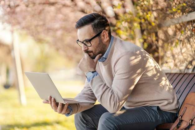 Zamyślony mężczyzna siedzi w parku i używa laptopa