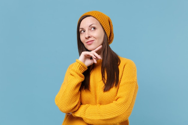 Zamyślony młoda brunetka kobieta dziewczyna w żółty sweter i kapelusz pozowanie na białym tle na niebieskim tle portret studio. Koncepcja życia szczere emocje ludzi. Makieta miejsca na kopię. Umieść podpórkę na podbródku.