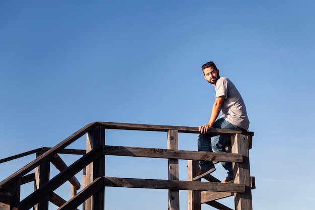 Zamyślony młody człowiek siedzący na drewnianej balustradzie z błękitnym niebem w tle koncepcja wypoczynku i relaksu kopia miejsca na tekst