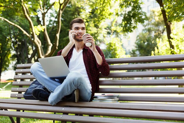 Zamyślony młody mężczyzna rozmawia przez telefon komórkowy, pracuje na laptopie i pije kawę, siedząc w parku na ławce. Koncepcja technologii, komunikacji, edukacji i pracy zdalnej, kopia przestrzeń