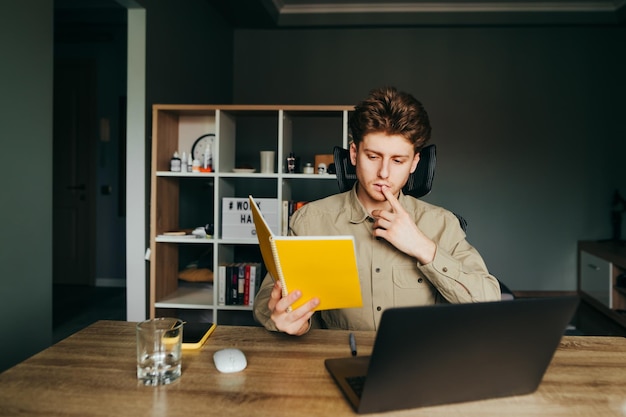 Zamyślony student studiuje zdalnie w domu, siedzi w pracy z laptopem i notatnikiem w ręku, czyta i myśli, że Guy uczy się online w kwarantannie