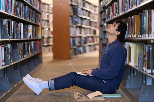 Zamyślony, wielozadaniowy student oparty na półce z literaturą za pomocą laptopa w kampusie biblioteki