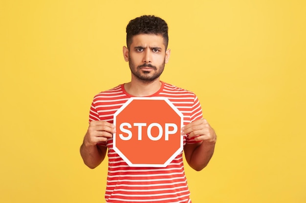 Zaniepokojony zły człowiek z brodą w paski tshirt trzymając czerwony znak stop ostrzeżenie o ograniczeniu prędkości niebezpieczna droga kryty strzał studio na białym tle na żółtym tle