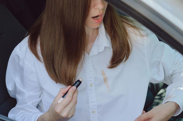 Zaskoczona kobieta pokazująca kosmetyczną plamę na białych ubraniach siedząc w samochodzie