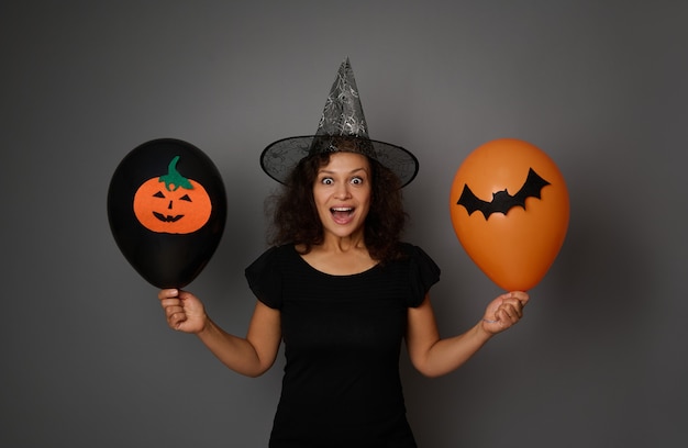 Zaskoczona kobieta w kapeluszu czarodzieja pozuje z czarno-pomarańczowymi balonami z wyciętym filcem nietoperzem i dynią, patrząc w kamerę. Koncepcja Halloween na szarym tle, skopiuj miejsce na reklamę