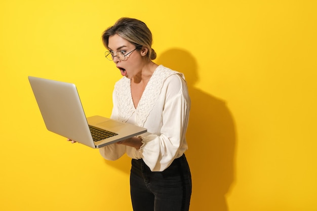 Zaskoczona kobieta w okularach, korzystająca z laptopa na żółtym tle