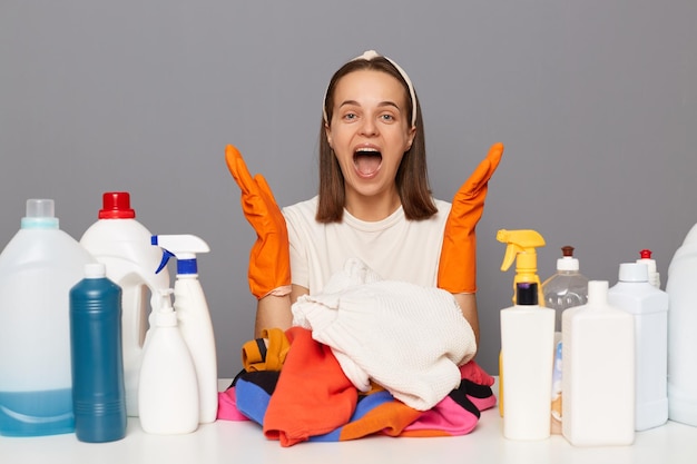 Zaskoczona kobieta w pomarańczowych gumowych rękawiczkach pozuje w miejscu pracy z detergentami czyszczącymi odizolowanymi na szarym tle, krzycząc ze szczęścia, ciesząc się pracą w domu