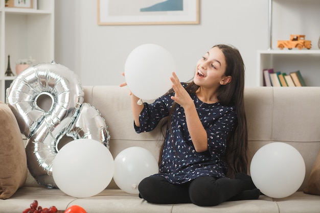 Zaskoczona mała dziewczynka w szczęśliwy dzień kobiety, trzymająca i patrząca na balon siedzący na kanapie w salonie