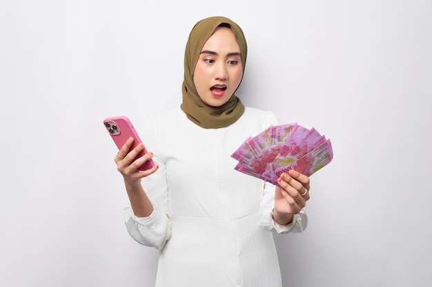 Zaskoczona młoda arabska azjatycka muzułmanka lat 20. nosząca hidżab trzymająca gotówkę w banknocie rupii i telefon komórkowy izolowana na białym tle Koncepcja religijnego stylu życia ludzi