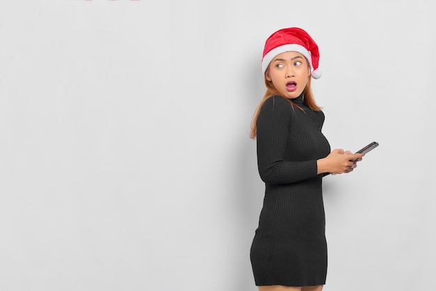 Zaskoczona młoda Azjatka w kapeluszu Świętego Mikołaja trzymająca telefon komórkowy i odwracająca wzrok na miejsce na kopię na białym tle nad białym tłem