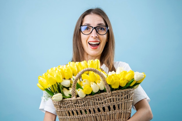 Zaskoczona młoda kobieta w okularach trzymająca żółte tulipany w koszu na niebieskim tle