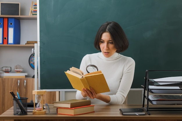 zaskoczona młoda nauczycielka patrząca na książkę z lupą siedzącą przy biurku z włączonymi narzędziami szkolnymi w klasie