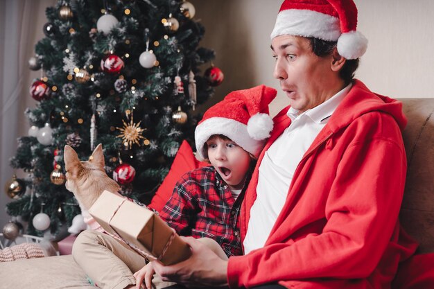 Zaskoczona Rodzina W Santa Hat Ojciec I Syn Otwierają Prezent świąteczny W Domu Siedząc Na Kanapie In