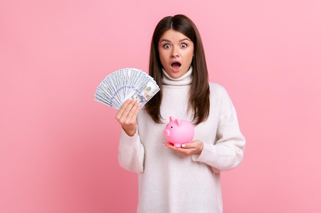 Zaskoczony brunetka kobieta trzyma fan banknotów dolarowych i wzrost oszczędności depozytów na sobie biały sweter w stylu casual Kryty studio strzał na białym tle na różowym tle