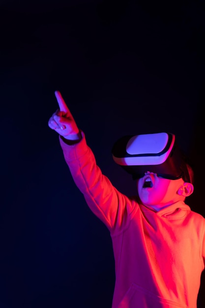 Zdjęcie zaskoczony chłopiec wskazujący w górę w goglach wirtualnej rzeczywistości oświetlonych futurystycznymi neonami