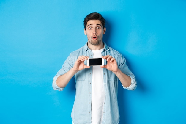 Zaskoczony facet pokazujący ekran telefonu komórkowego i wyglądający na pod wrażeniem, stojący na niebieskim tle