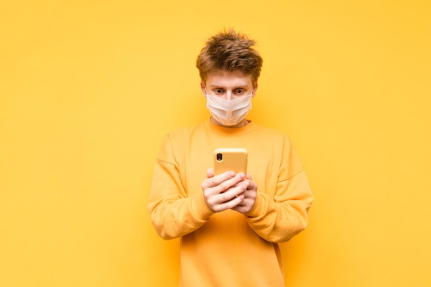 Zaskoczony facet w masce medycznej i żółtej bluzie korzysta z internetu na swoim smartfonie