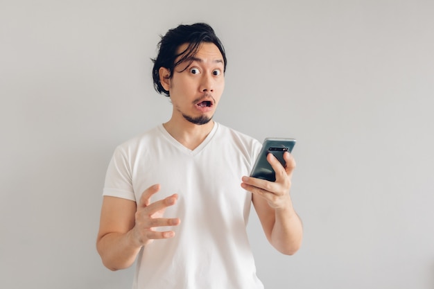 Zaskoczony i zszokowany długowłosy mężczyzna w białej koszulce używa smartfona.