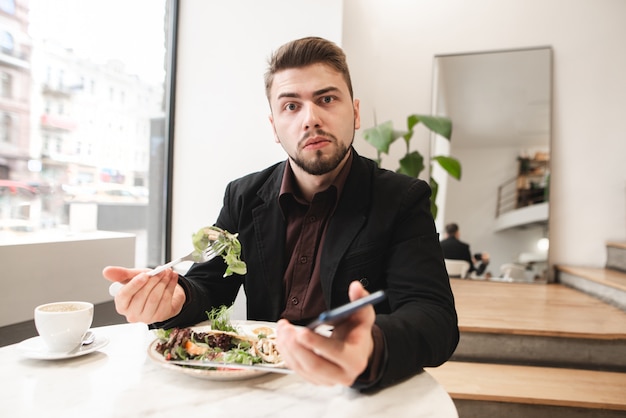 Zaskoczony mężczyzna w garniturze siedzi w przytulnej restauracji ze smartfonem w rękach, je sałatkę z talerzem z widelcem i patrzy w kamerę.