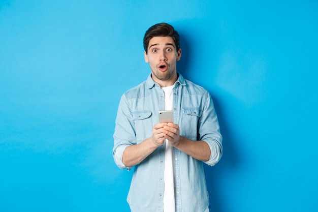 Zaskoczony mężczyzna w zwykłym ubraniu wyglądający na zdumionego, trzymający smartfon, stojący przed niebieską ścianą