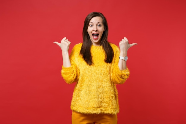 Zaskoczony młoda kobieta w żółtym futerkowym swetrze, trzymając otwarte usta, wskazując kciuki na bok na białym tle na jasnoczerwonym tle ściany w studio. Ludzie szczere emocje, koncepcja stylu życia. Makieta miejsca na kopię.