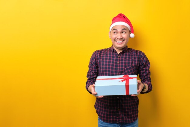 Zaskoczony młody Azjata w czapce Świętego Mikołaja trzymający pudełko ze wstążką, patrzący na bok na miejsce kopiowania na żółtym tle studia Szczęśliwego Nowego Roku 2023 celebracja wesołych świąt koncepcja