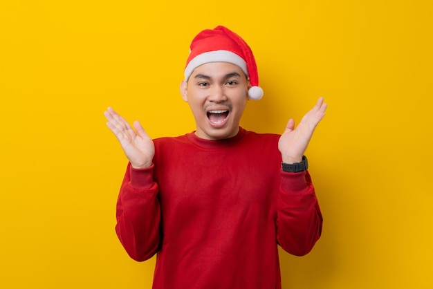 Zaskoczony młody Azjata w kapeluszu Świętego Mikołaja podnoszący ręce i patrzący na kamerę z wyrazem podekscytowania na żółtym tle studia obchody świąt Bożego Narodzenia i koncepcja Nowego Roku
