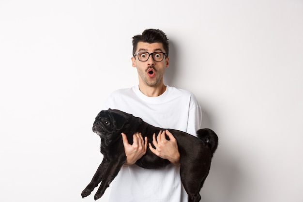 Zaskoczony młody mężczyzna w okularach trzymający ślicznego czarnego mopsa, właściciel psa wpatrzony w kamerę z wyrazem twarzy, mówiący wow, stojąc na białym.