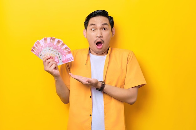 Zaskoczony młody przystojny azjatycki mężczyzna pokazujący banknoty pieniężne na żółtym tle