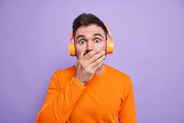 Zaskoczony przystojny mężczyzna zakrywa usta i wygląda na oszołomionego, będąc zszokowany czymś strasznym, ma na sobie jasnopomarańczowy sweter i słucha ścieżki dźwiękowej przez bezprzewodowe słuchawki