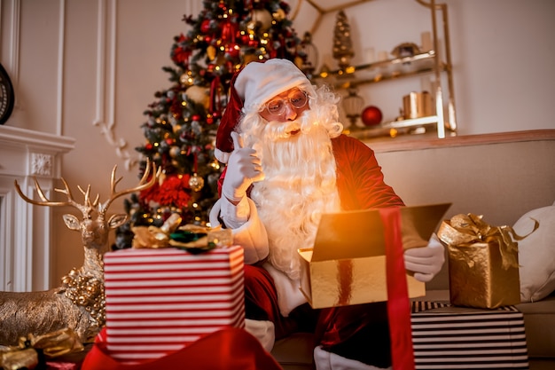 Zaskoczony Święty Mikołaj z magicznym świecącym prezentem w pobliżu pięknej choinki. Nowy rok i Wesołych Świąt, koncepcja wesołych świąt