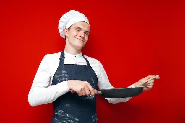 Zaskoczony szef kuchni trzyma naczynia i przygotowuje danie na czerwonym, izolowanym tle pracownik kuchni w mundurze z przedmiotami kuchennymi
