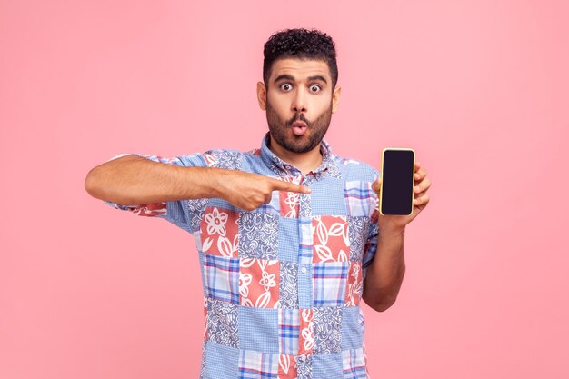 Zaskoczony zdezorientowany mężczyzna z brodą na sobie niebieską koszulę wskazującym palcem na smartfonie z czarnym pustym ekranem kopią miejsca na reklamę Kryty studio strzał na białym tle na różowym tle