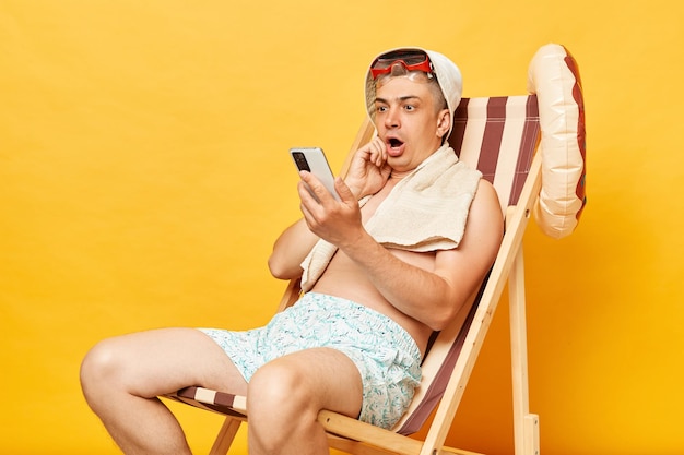 Zaskoczony zdumiony zdumiony mężczyzna bez koszuli siedzący na krześle izolowany na żółtym tle używający telefonu komórkowego z zszokowanym wyrazem twarzy czytający nieprzyjemne wiadomości