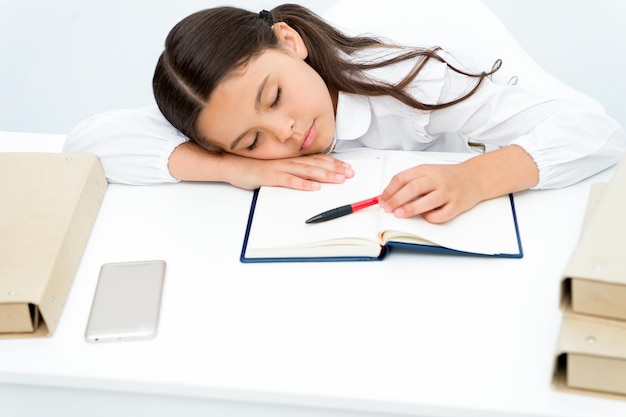 Zasnąć na lekcji. Dziecko dziewczynka zasnąć podczas czytania książki stół białe tło. Uczennica zmęczona studiowaniem i czytaniem książki. Dziecko dziewczynka szkolny mundurek senna twarz leżała na otwartej księdze i spać.