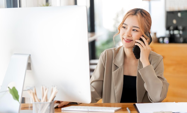 Zastrzelona szczęśliwa businesswoman siedząca przy biurku pracująca na komputerze stacjonarnym i rozmawiająca z kimś na swoim telefonie komórkowym podczas pracy w domu Biuro w domu